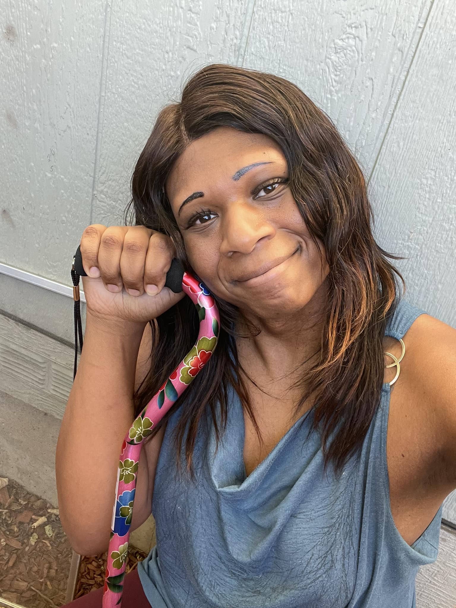 Picture of Hannah - a black trans woman crouching on a concrete porch and leaning against a green colored wall of house.  She has long black hair, a teal shirt with a picture of a wolf and is smiling.  She is proudly holding a cane in her right hand.