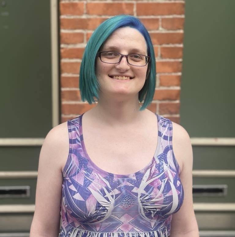 Picture of Josie - a smiling blue-haired trans woman with purple-rimmed glasses standing in front of a brick wall